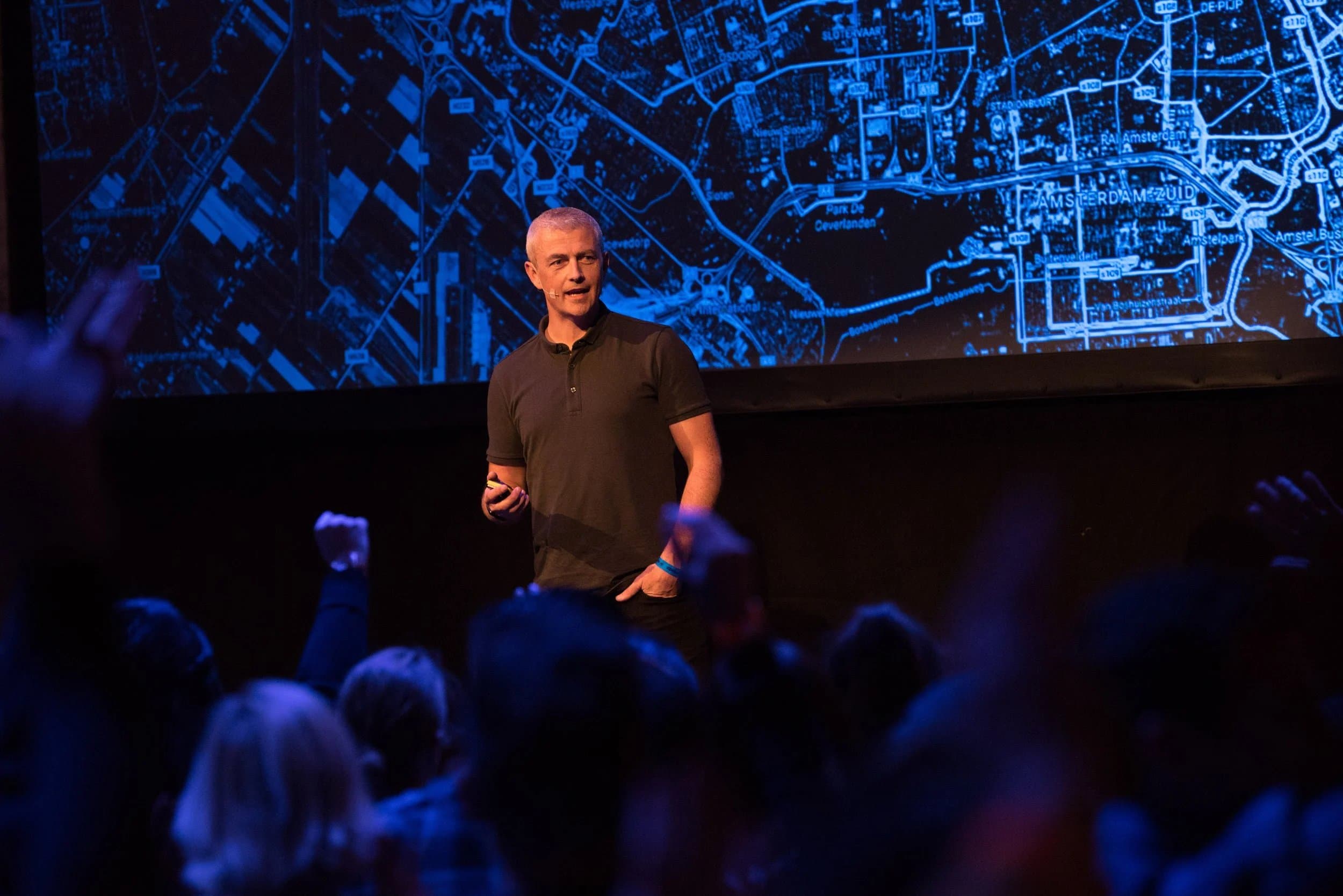 Scott Robertson speaking on stage in front of a projected map of Amsterdam