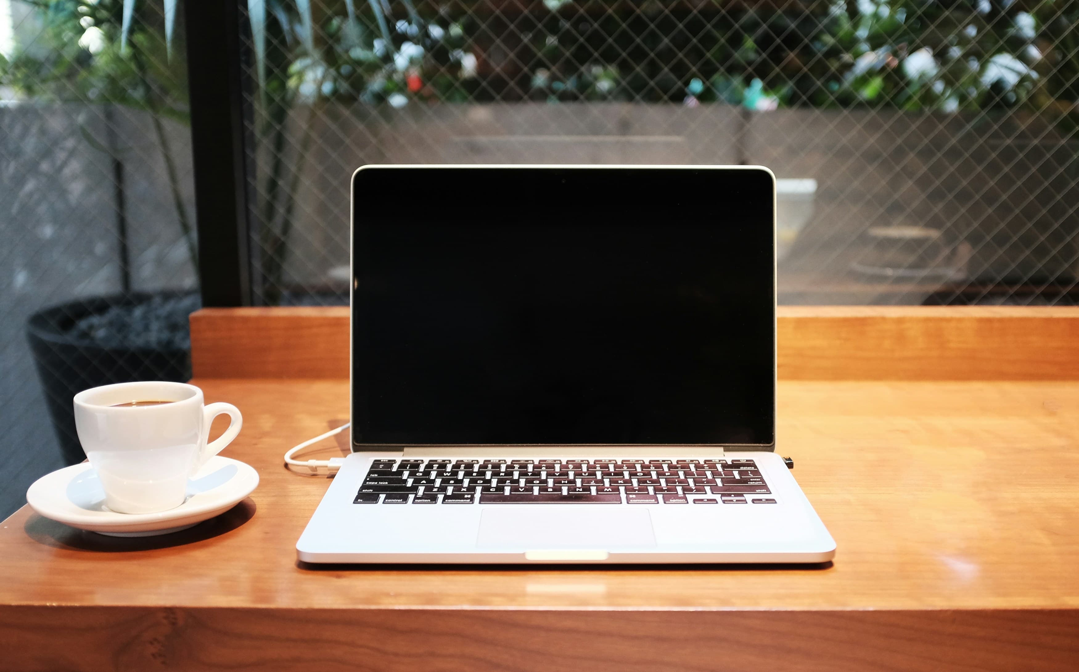 A silver laptop and a white espresso cup on a saucer, on a wooden café table by a window.