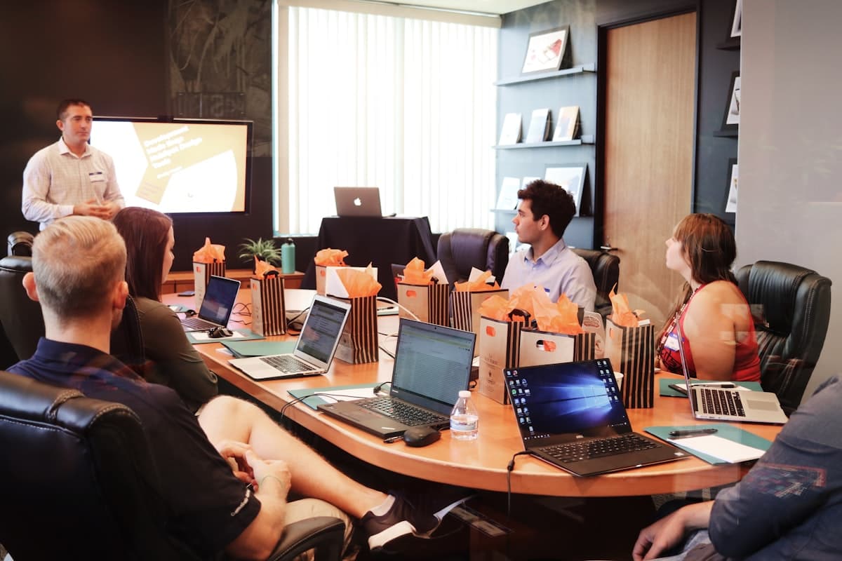 A team meeting around a boardroom table with laptops open, a presenter addressing the group